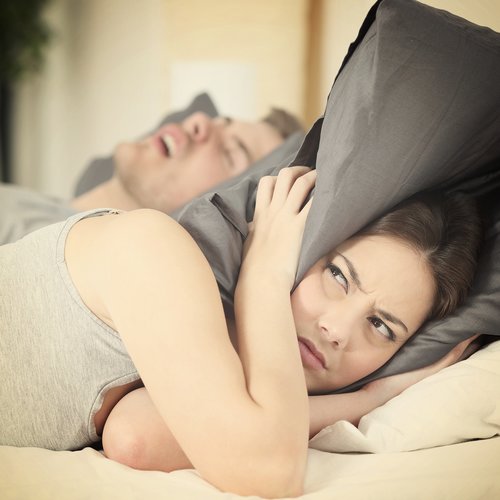 Young woman laying in bed with her boyfriend with pillow around her ears because she can't sleep